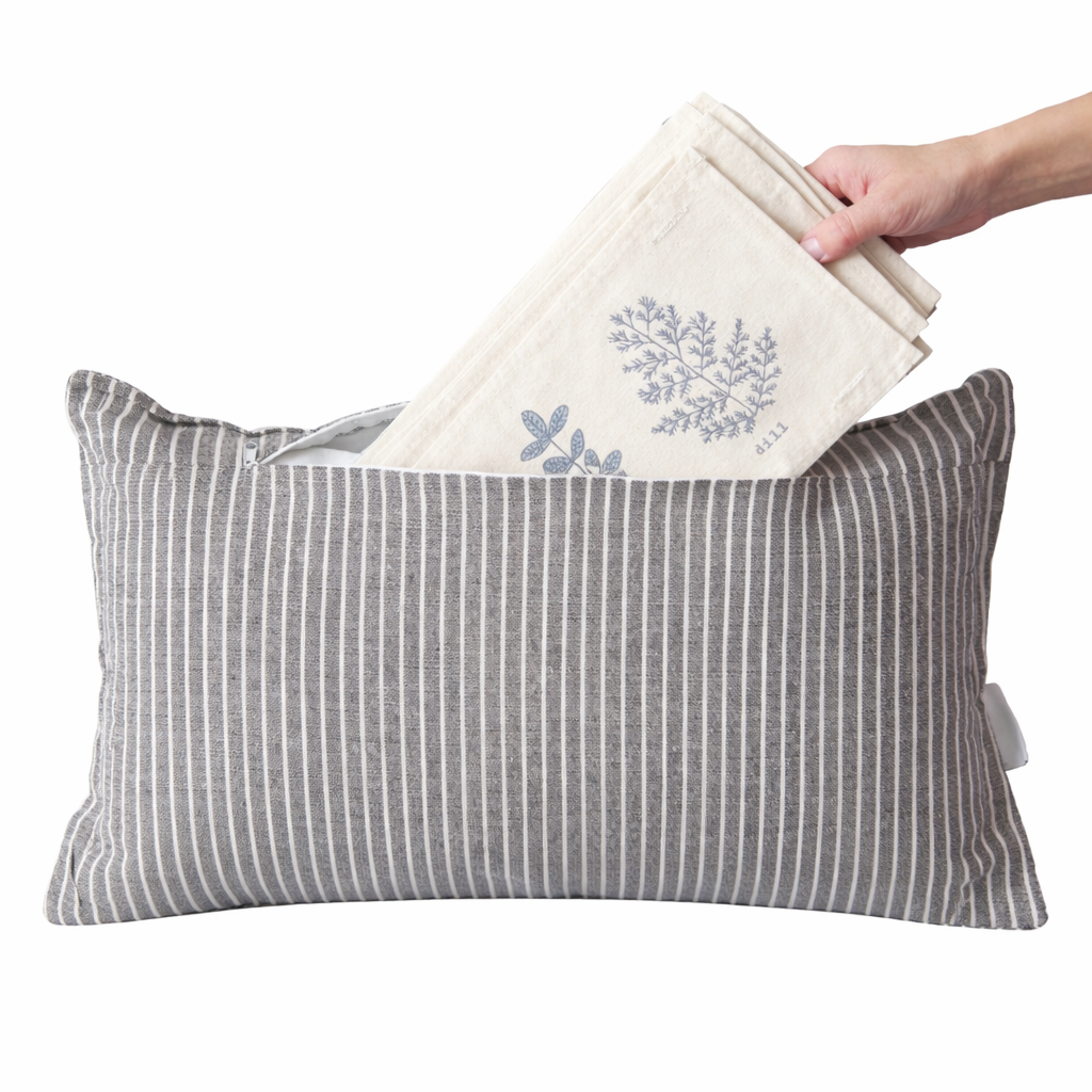A gray and white vertically striped rectangular pillow with a hand holding a folded cream linen cloth with a blue botanical print above it, against a plain white background.