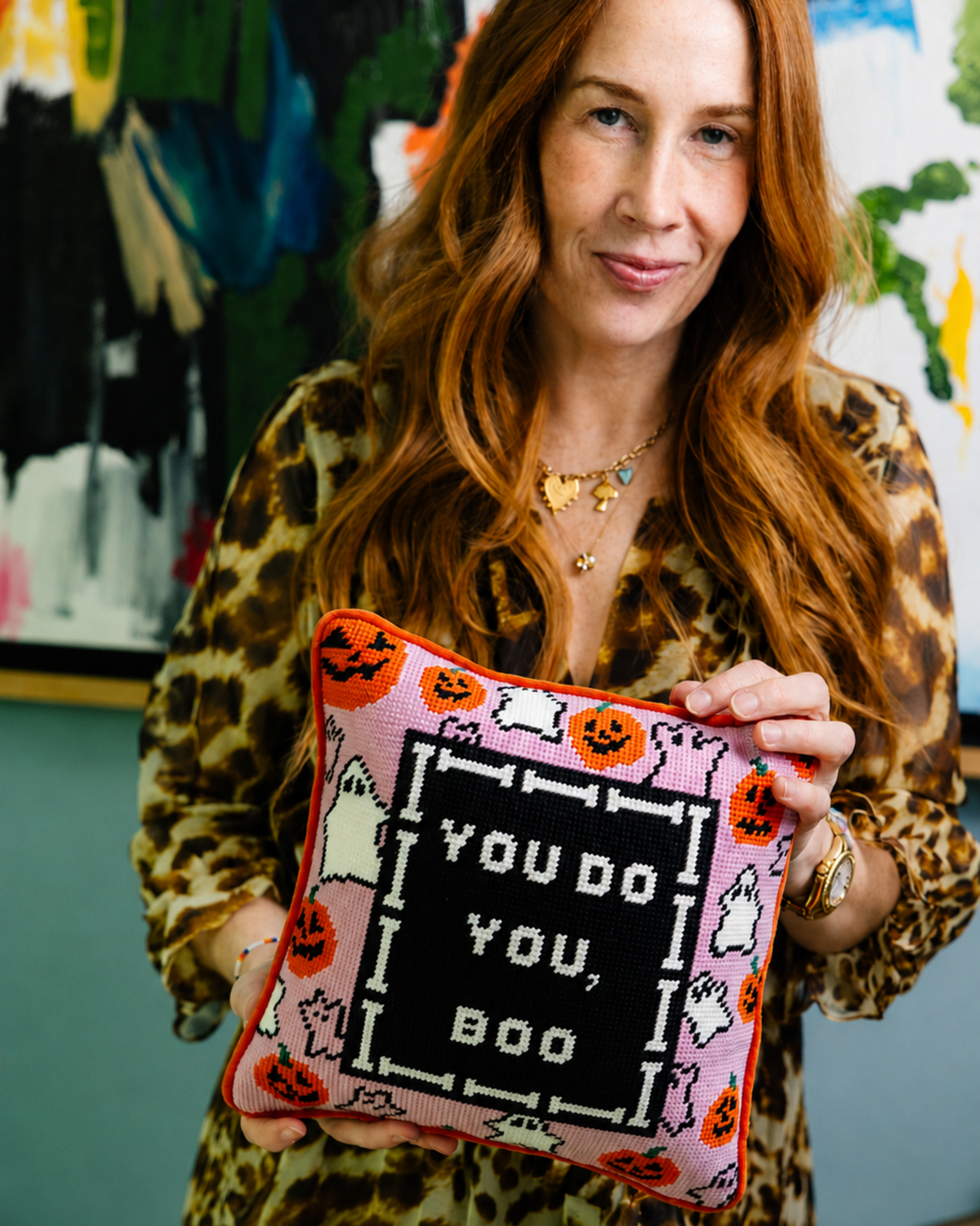 A woman with long red hair wearing a leopard print top holds up a square pink and black embroidered pillow with a Halloween theme and the text 'YOU DO YOU, BOO'.