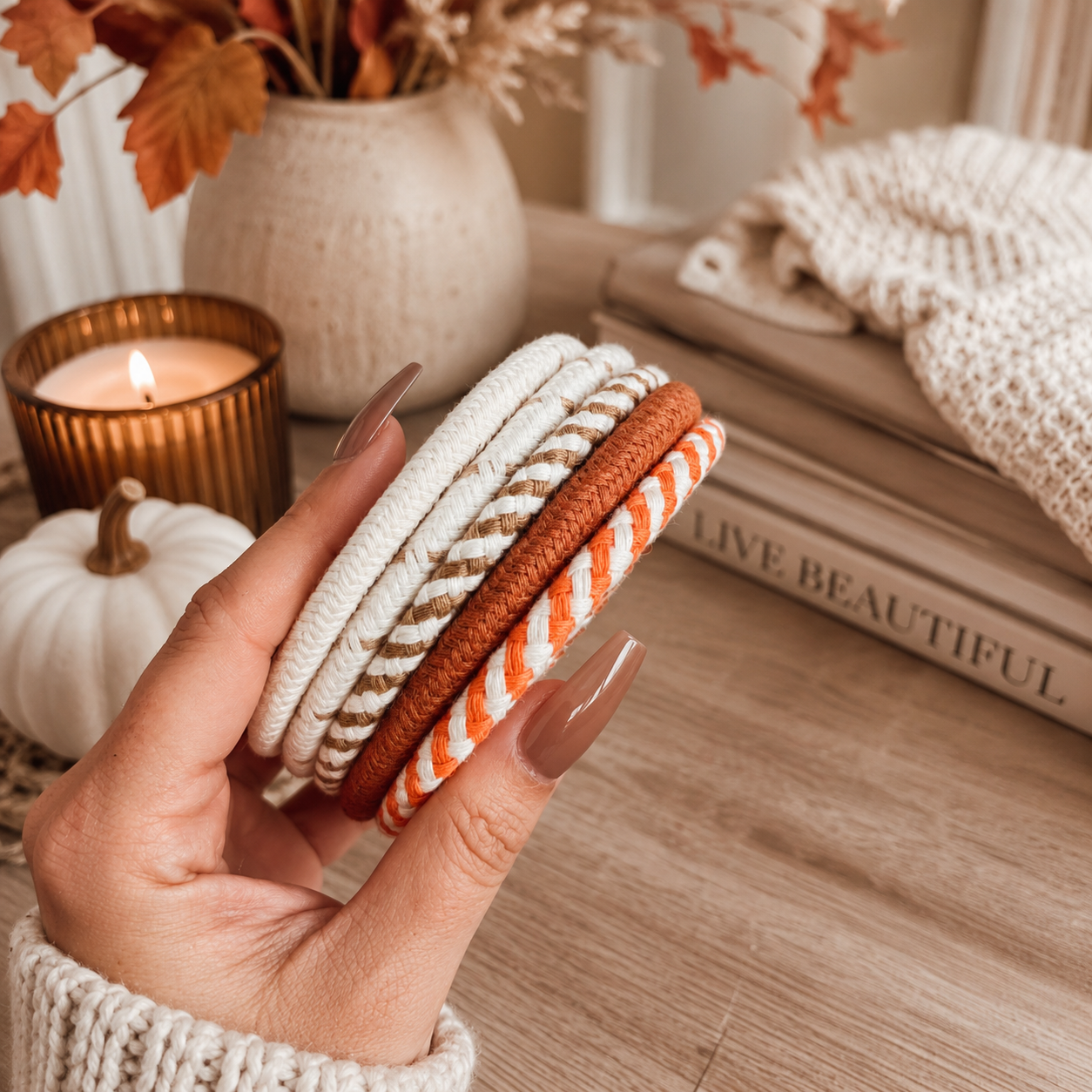 Hand holding three woven coasters with a cozy autumn setting featuring a candle, pumpkin, and books.