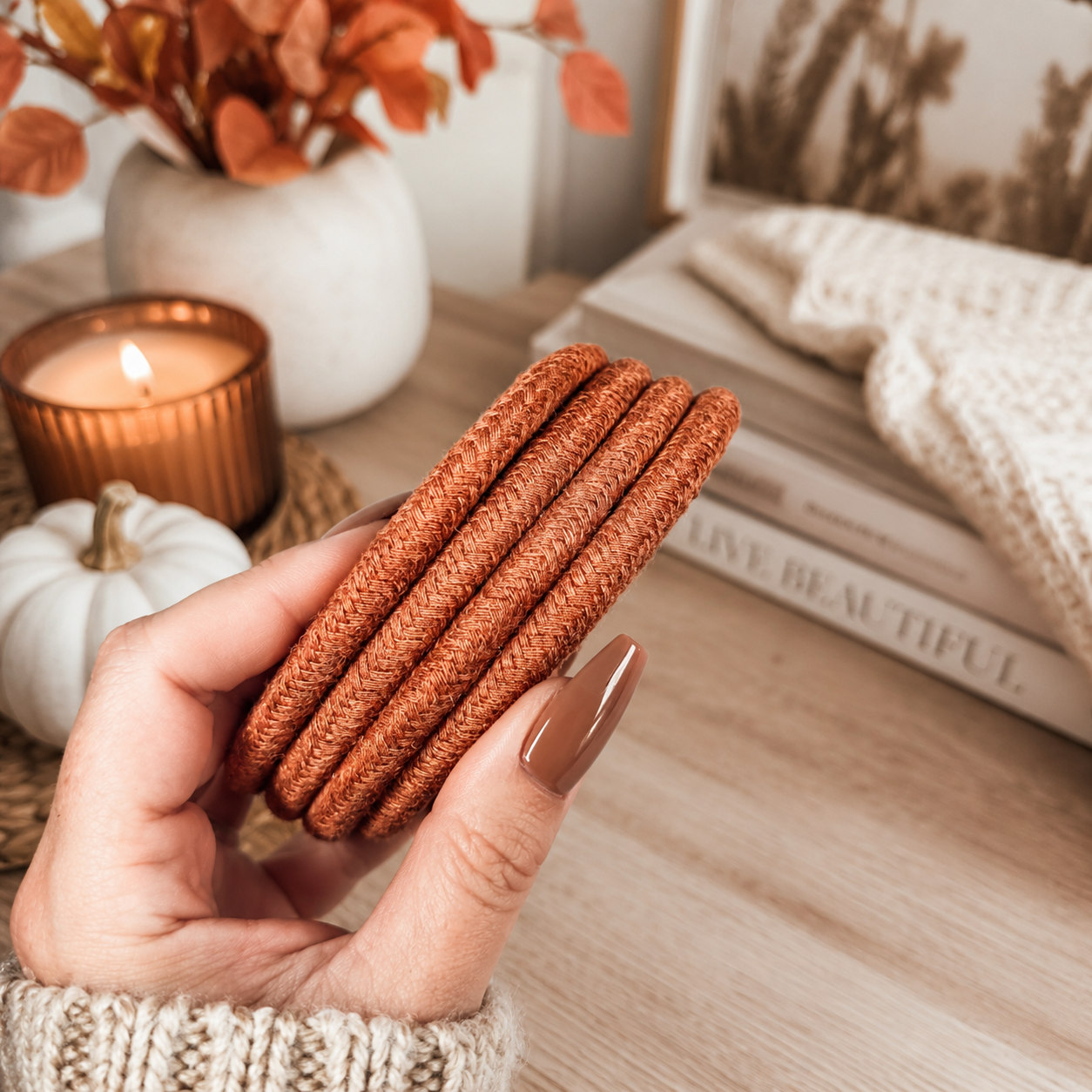 Hand holding brown coasters with a cozy indoor setting featuring a candle, books, and decorative items.