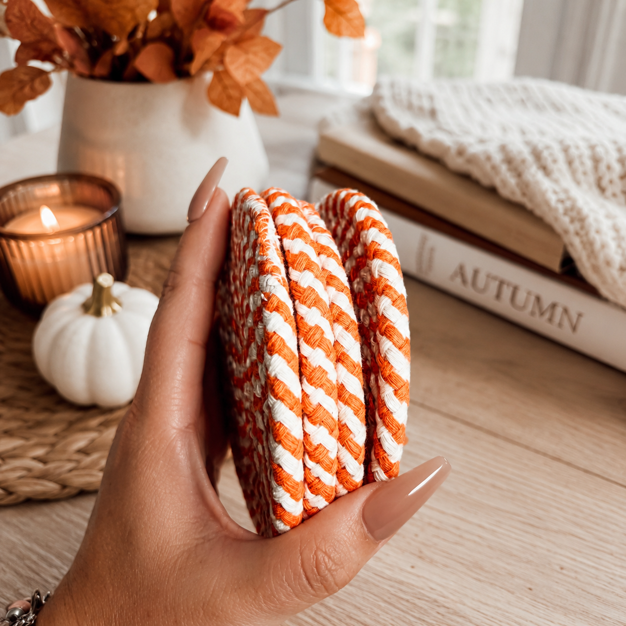 Hand holding orange and white striped coasters with a cozy autumn setting in the background.