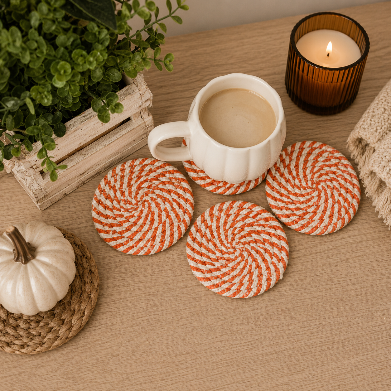 White mug with coffee on orange and white striped coasters on a wooden table with a candle and plant.