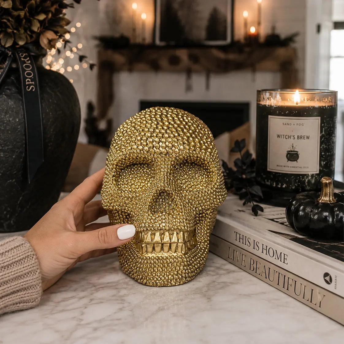 Gold skull decorative item held by a hand with a candle and books in the background