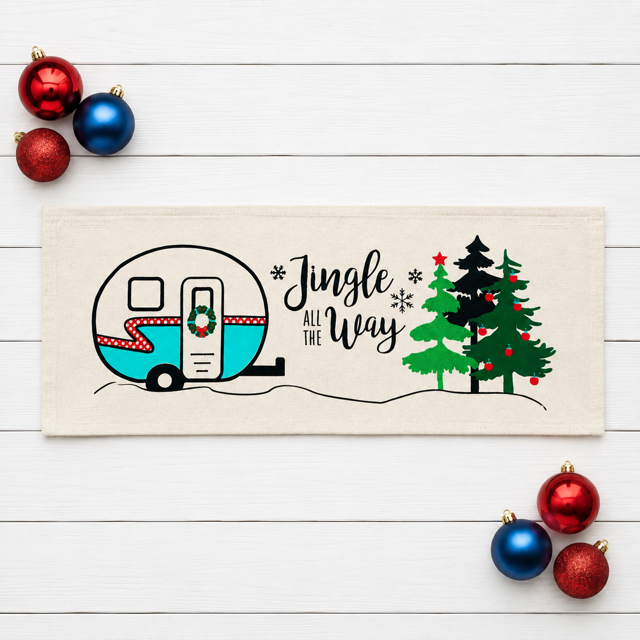 A rectangular cream-colored fabric tea towel or runner laid flat on a white wood background, decorated with a black line-art illustration of a retro camper, Christmas trees, snowflakes, and the phrase 'Jingle All The Way.' Red and blue ornaments are placed around it as styling props.