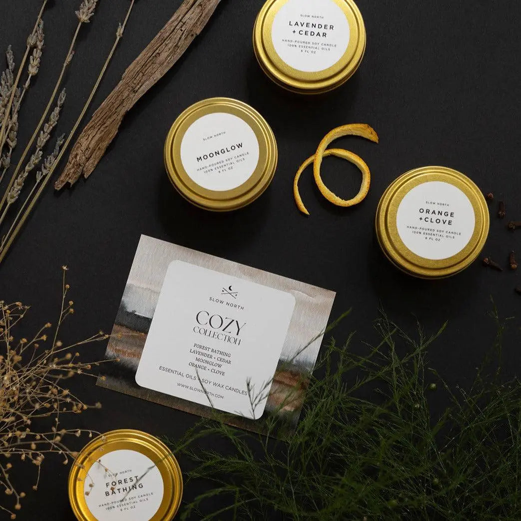 Cozy Collection soy candles in gold tins with dried botanicals, featuring Lavender+Cedar, Moonglow, Orange+Clove, and Forest Bathing scents
