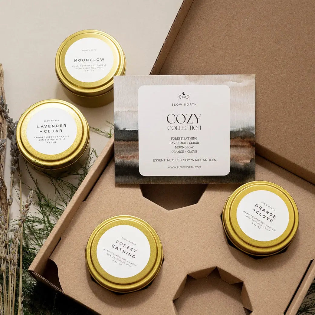 Cozy Collection soy wax candles with essential oils in gold tins, featuring Forest Bathing, Lavender + Cedar, Moonglow, and Orange + Clove scents