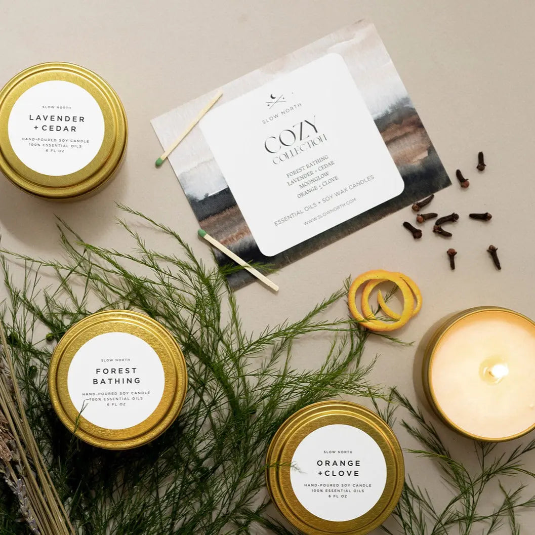 Cozy collection soy candles in gold tins with lavender, forest bathing, and orange clove scents, styled with greenery and cloves