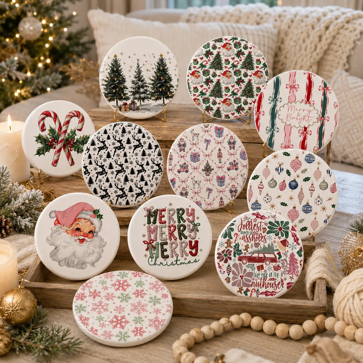 A set of round ceramic coasters with various Christmas-themed patterns, displayed on small stands on a wooden tray surrounded by holiday decorations like a candle, pine branches, beads, and a cozy knit blanket.
