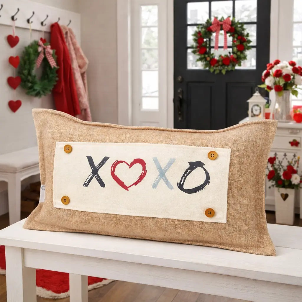 A rectangular burlap throw pillow with a white front panel featuring the words 'XOXO' in black, red, and light blue brushstrokes, with four wooden buttons on the sides, displayed on a white bench in a decorated holiday home.
