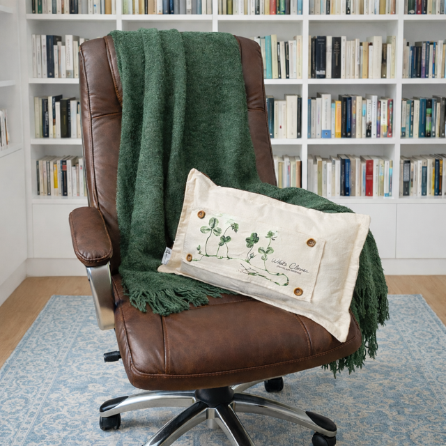 A brown leather office chair in a home office setting, styled with a dark green textured throw blanket and a cream-colored decorative lumbar pillow featuring green shamrock embroidery.
