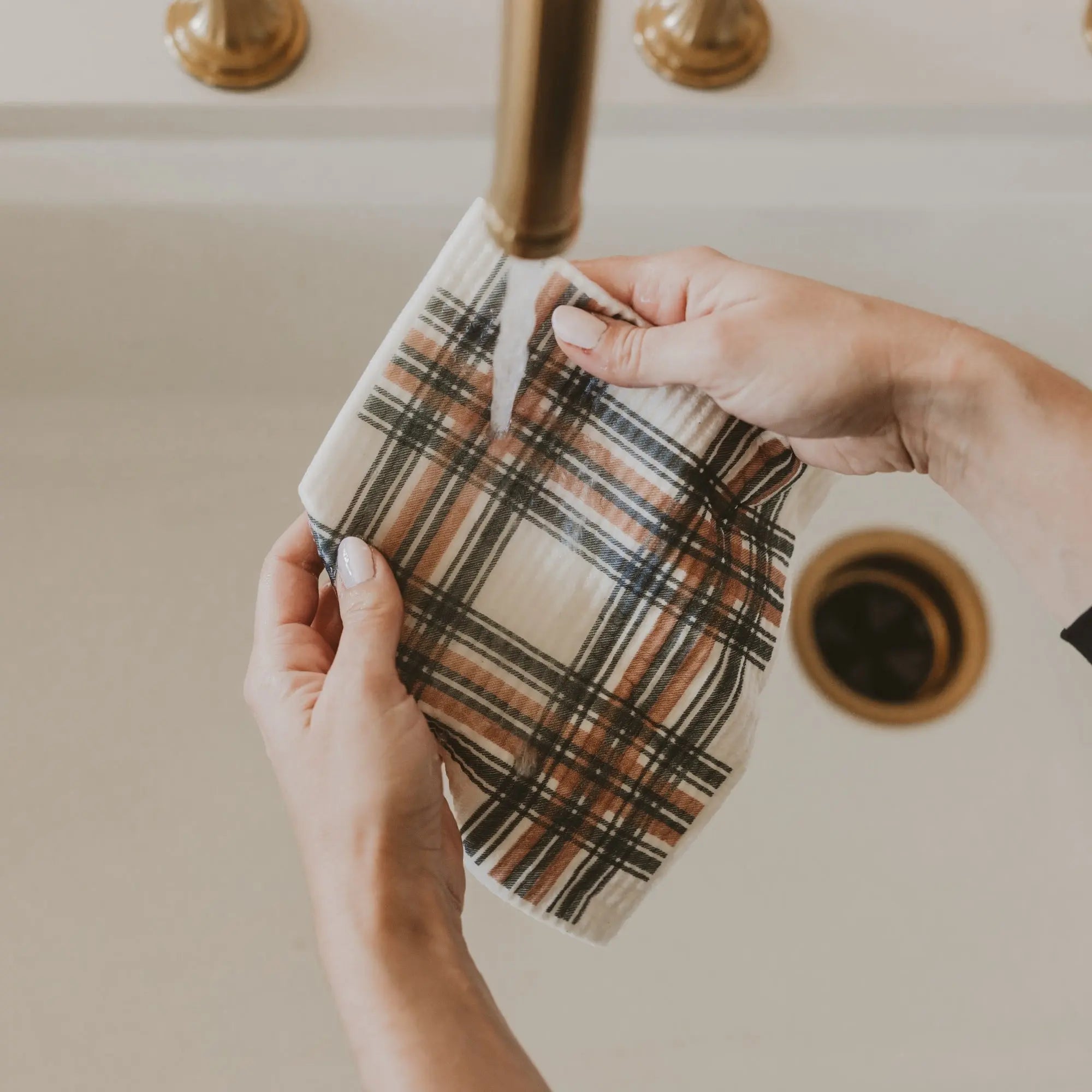 A person's hands holding a beige plaid kitchen towel under a brass faucet running in a sink.