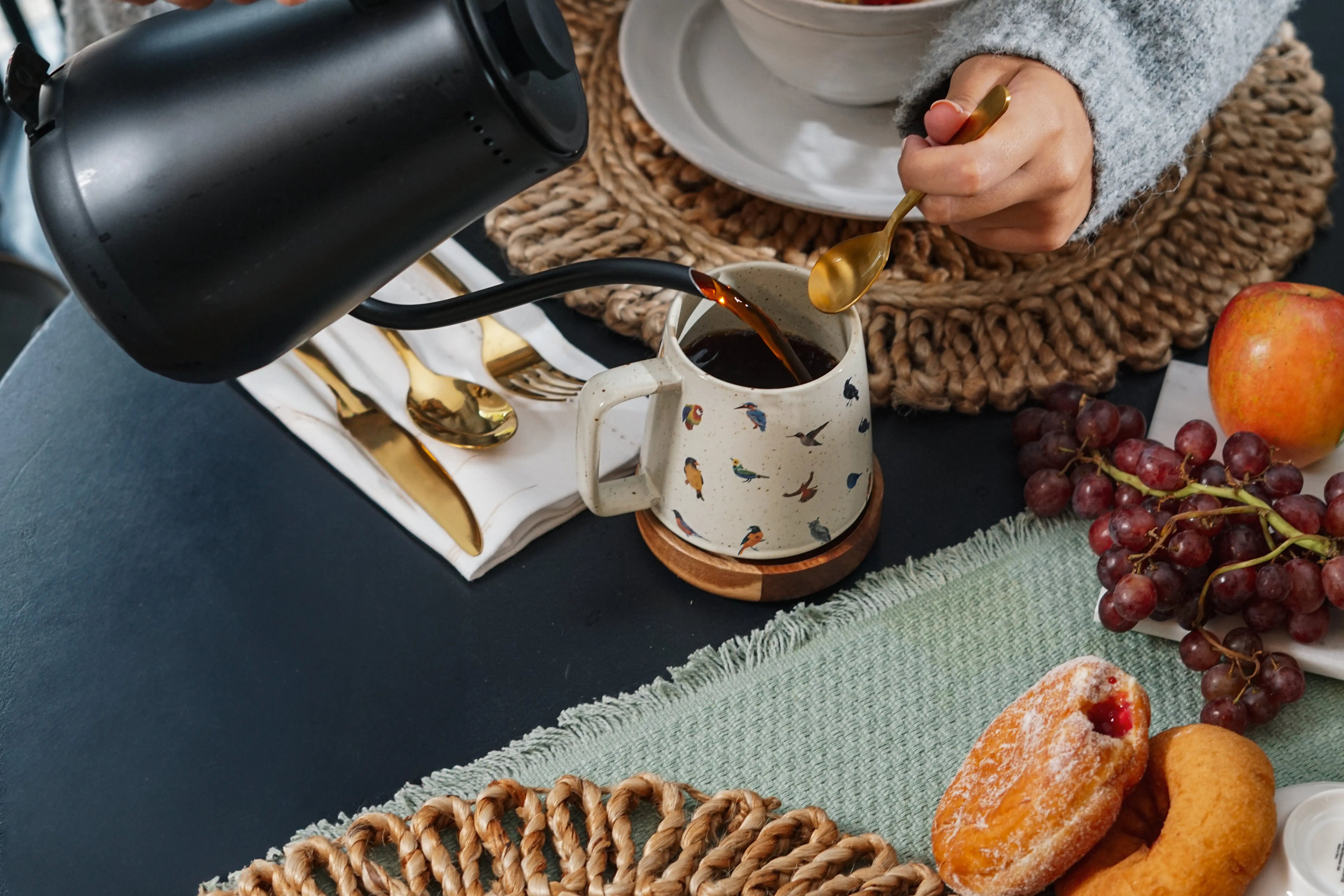Cozy farmhouse breakfast with coffee poured into bird-print mug, fresh grapes, apple, pastries, and woven placemats
