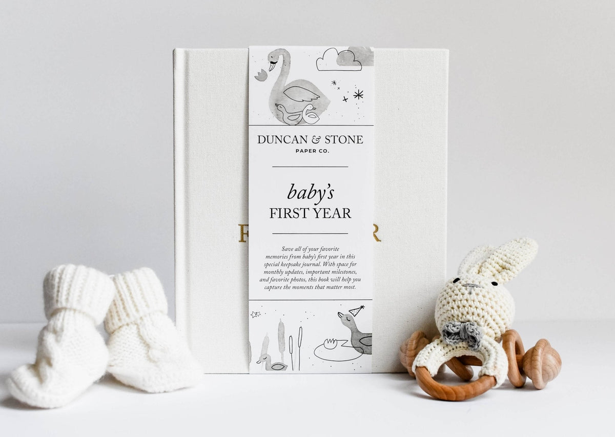 Book titled 'Baby's First Year' by Duncan & Stone with baby socks and a plush toy on a light gray background