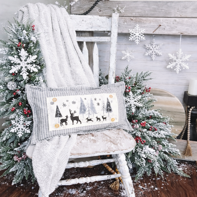 Decorative pillow with winter scene on a white chair surrounded by Christmas trees and snowflakes.