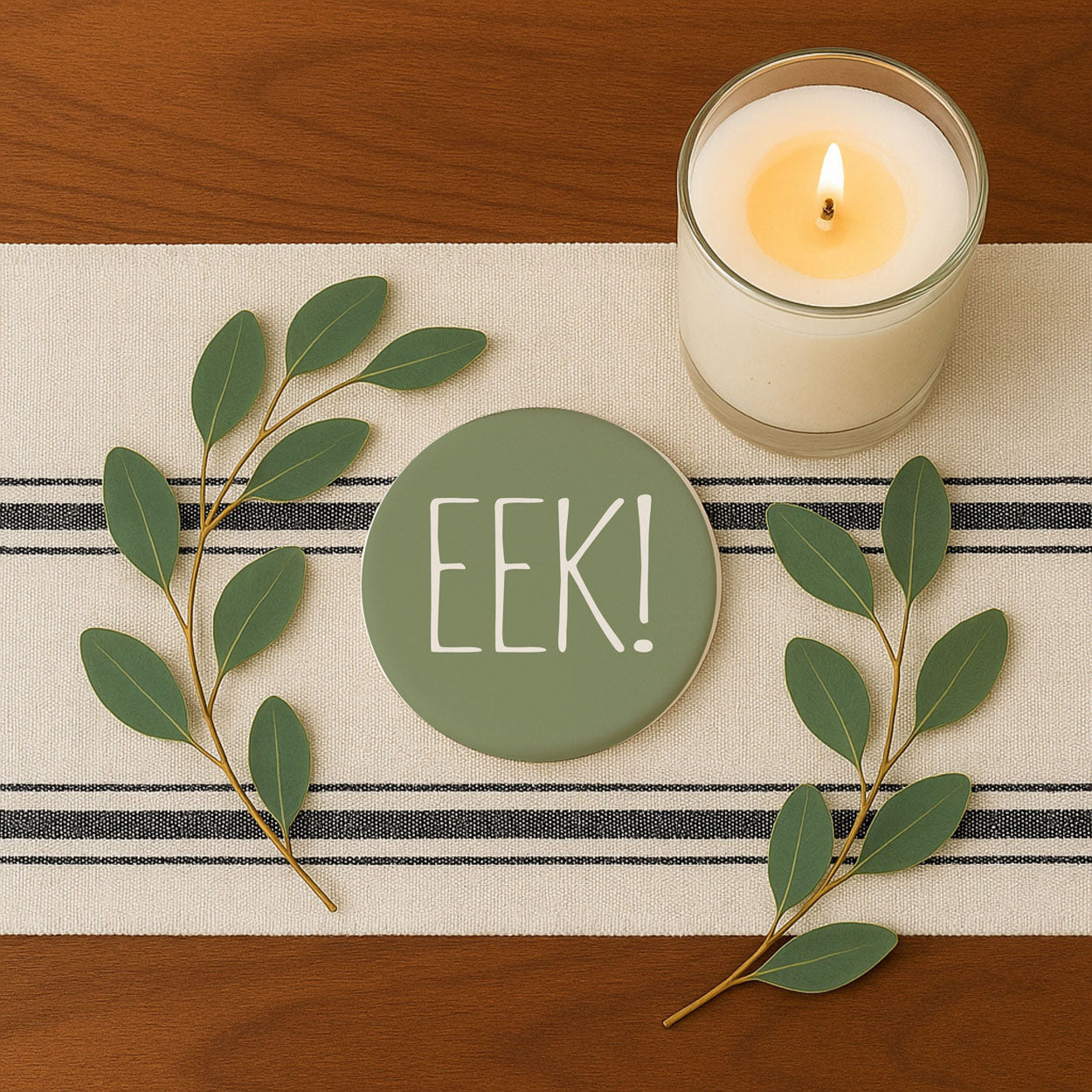 Candle with 'EEK!' coaster on a striped tablecloth with green leaves