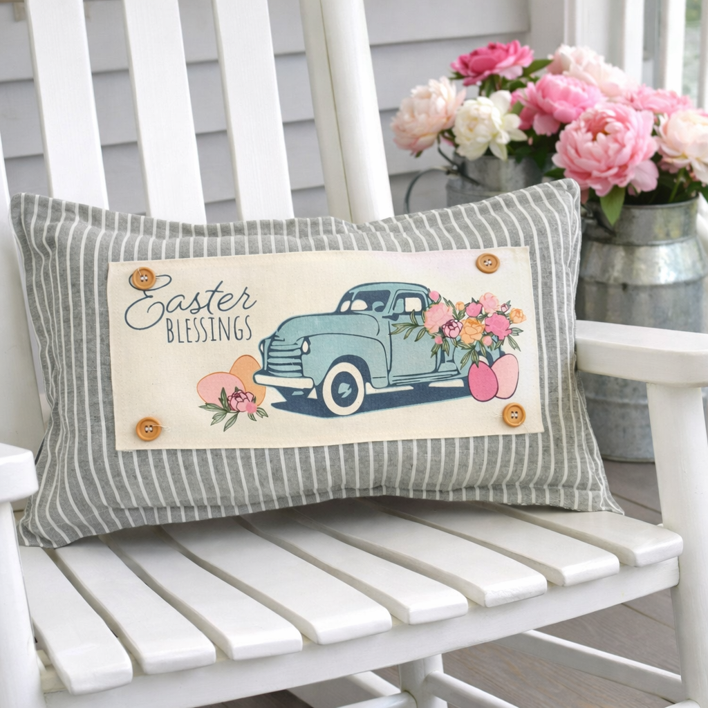 A rectangular decorative throw pillow with gray and white vertical stripes sits on a white wooden rocking chair. The center features a cream patch with a vintage light blue truck carrying pink and peach flowers, alongside the words 'Easter Blessings' and four decorative buttons.