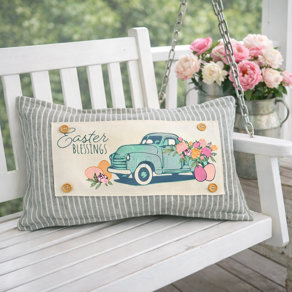 A rectangular decorative throw pillow with a gray and white striped fabric border and a central cream panel featuring a teal truck, flowers, and the words “Easter Blessings,” displayed on a white porch swing with pink flowers in the background.