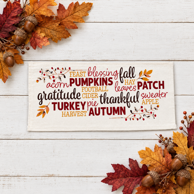 A rectangular cream fabric sign with autumn-themed typography and small leaf/berry illustrations, displayed on a white wood surface with fall leaves, acorns, and berries around it.
