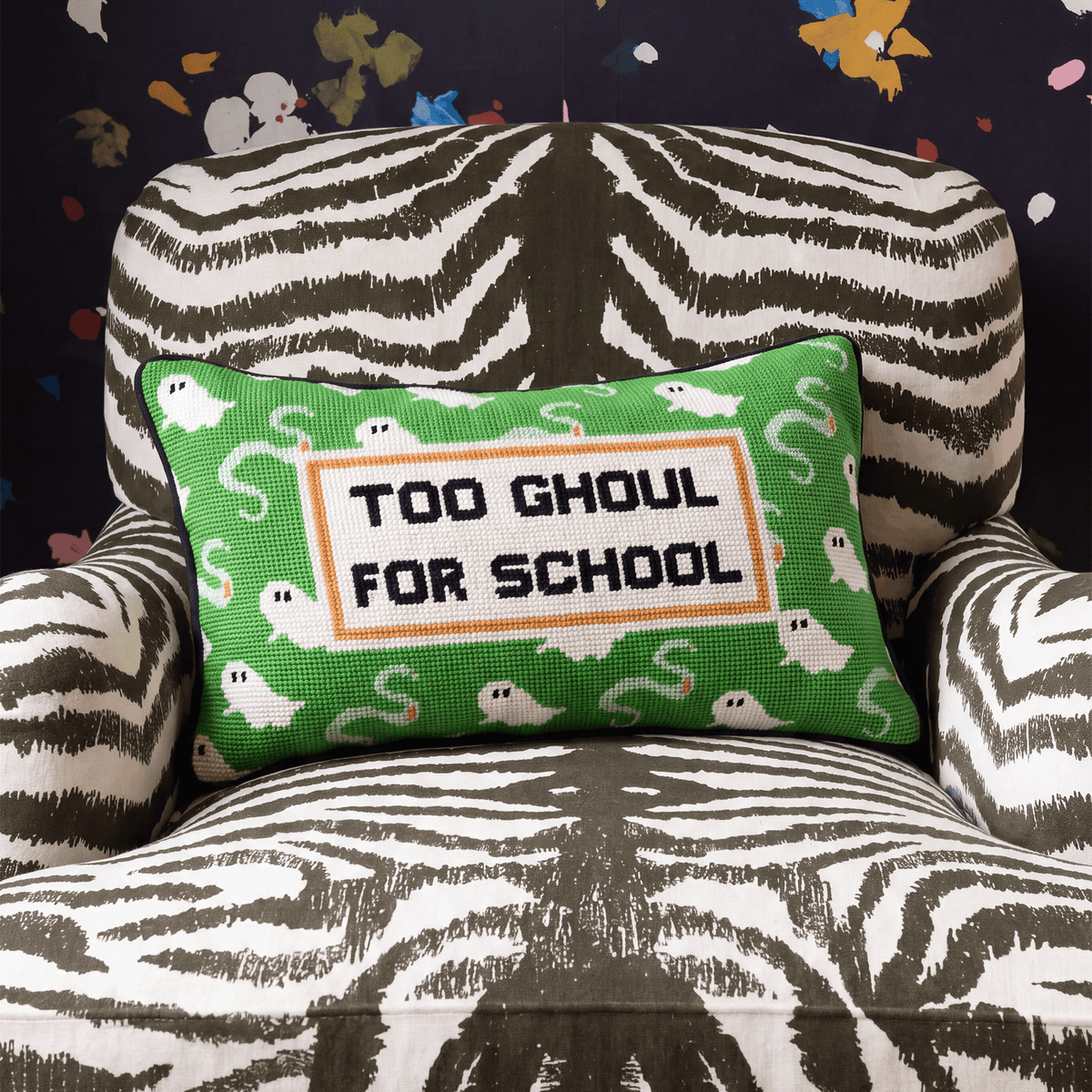A rectangular green throw pillow with a cross-stitch style design reading 'TOO GHOUL FOR SCHOOL' in black letters on a white panel, surrounded by small white ghosts and gray cobwebs, resting on a zebra-print armchair.
