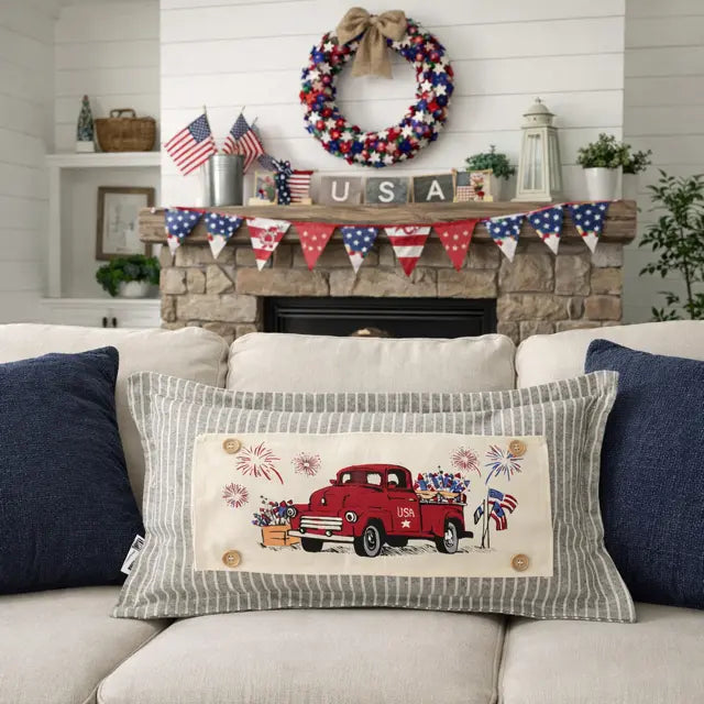 Decorative pillow with a red truck and American flag design on a couch, with a fireplace and festive decorations in the background.