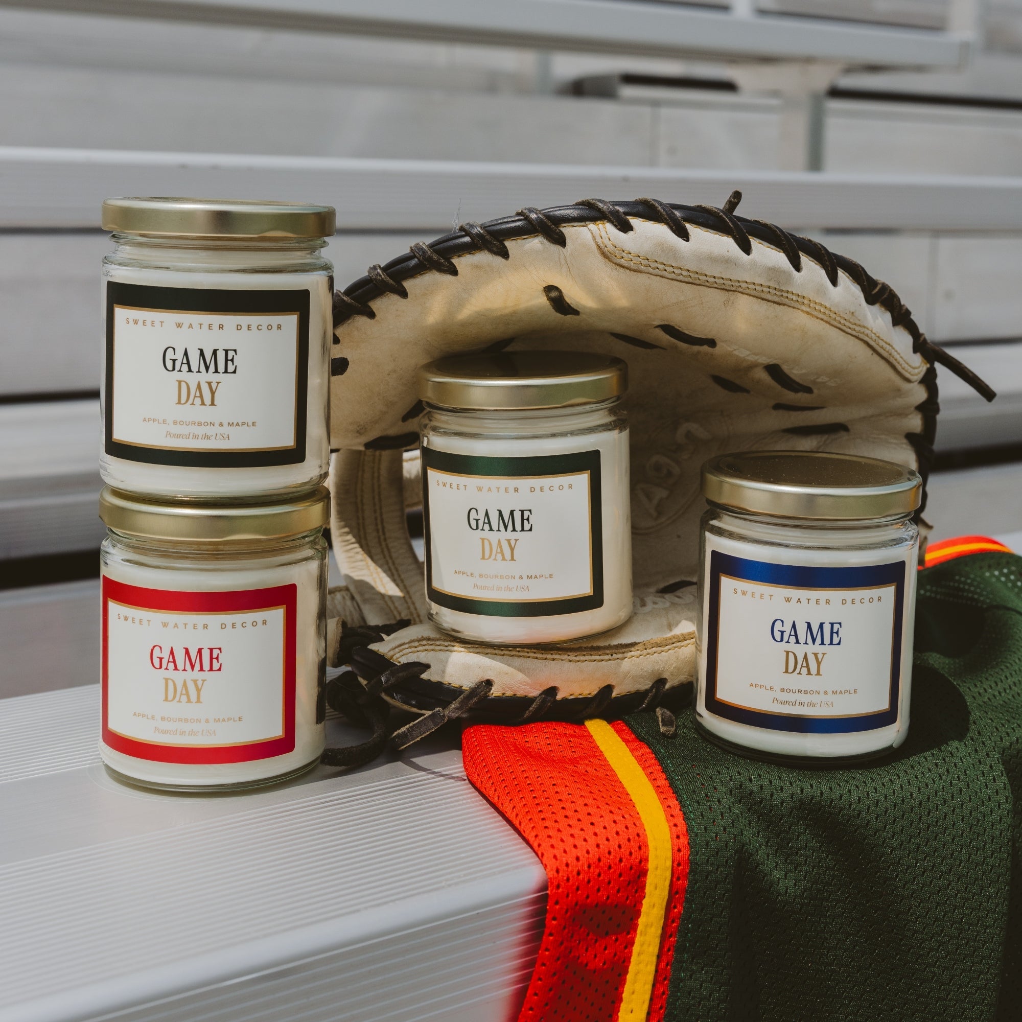 Four glass jar candles with gold lids and white labels reading 'GAME DAY' are arranged on a white bench with baseball-themed props, including a glove, a green jersey, and an orange-and-yellow strip.
