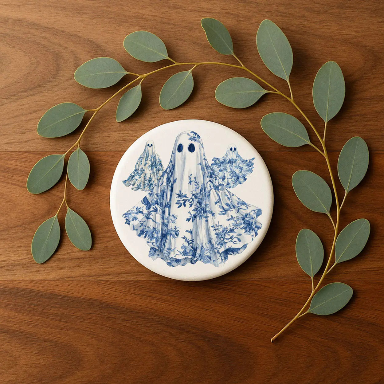 Round ceramic coaster with blue ghost design on a wooden surface with eucalyptus leaves.