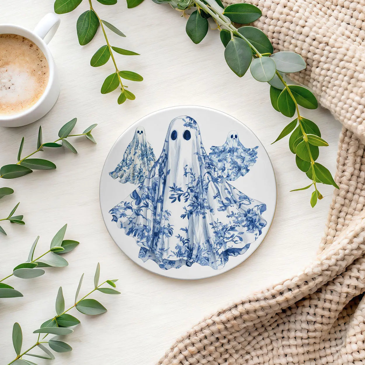 Round ceramic coaster with blue ghost design on a white surface with green leaves and a cup of coffee.