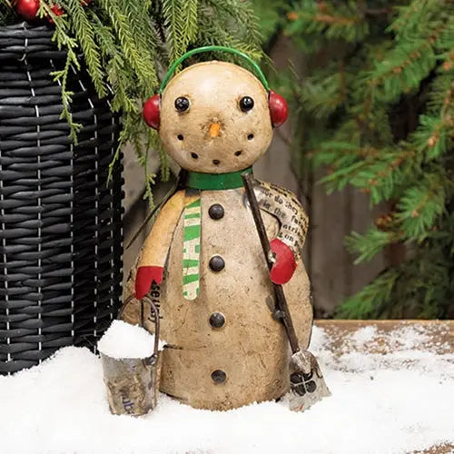 A rustic handmade snowman figurine made from a natural tree trunk, decorated with a green scarf, red earmuffs, and holding a small bucket and shovel, set in white faux snow with evergreen branches and a wicker basket in the background.