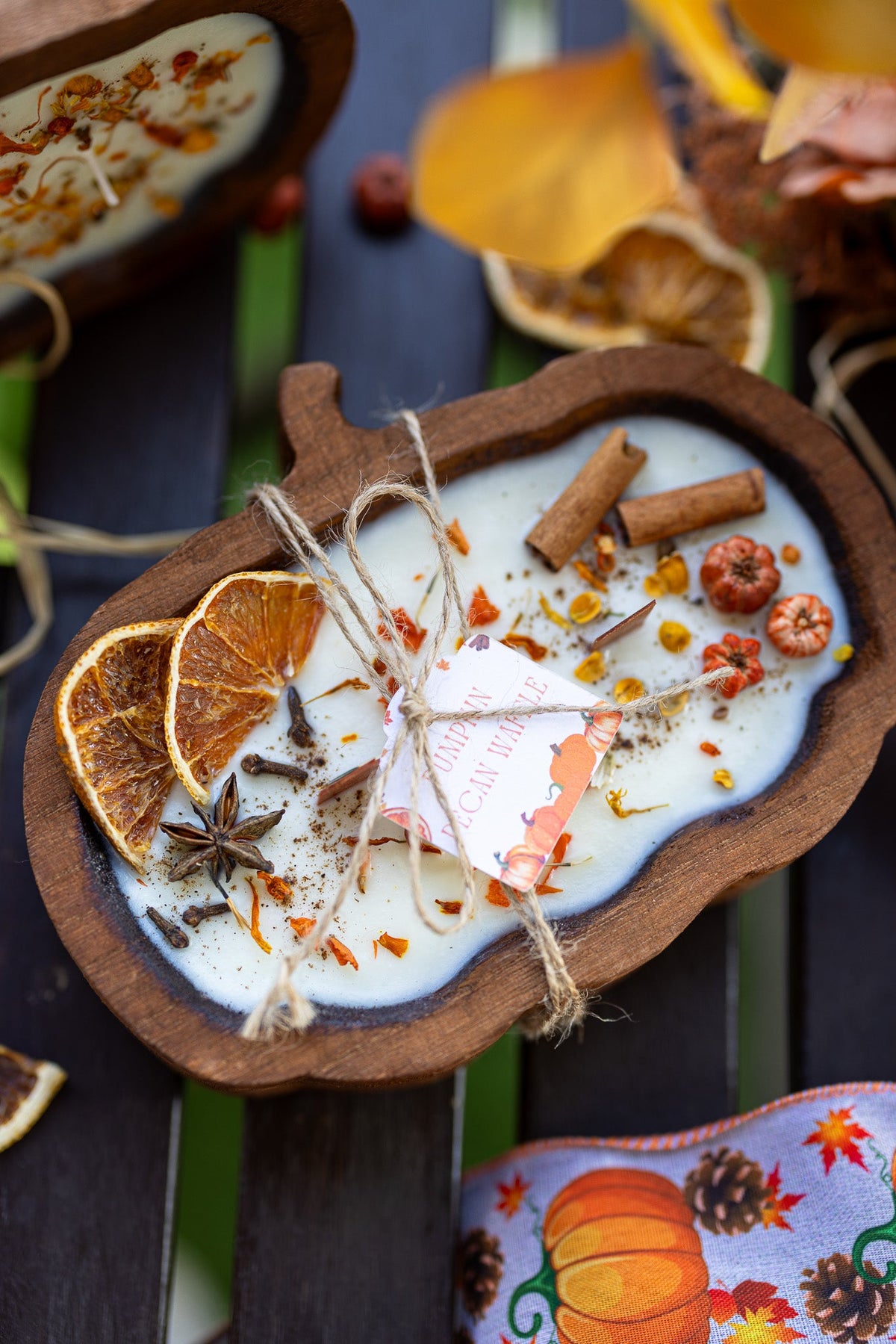 An autumn-themed wooden pumpkin-shaped candle in a dark rustic bowl, decorated with dried orange slices, star anise, cinnamon sticks, and a small hanging tag, set on a dark wooden surface.