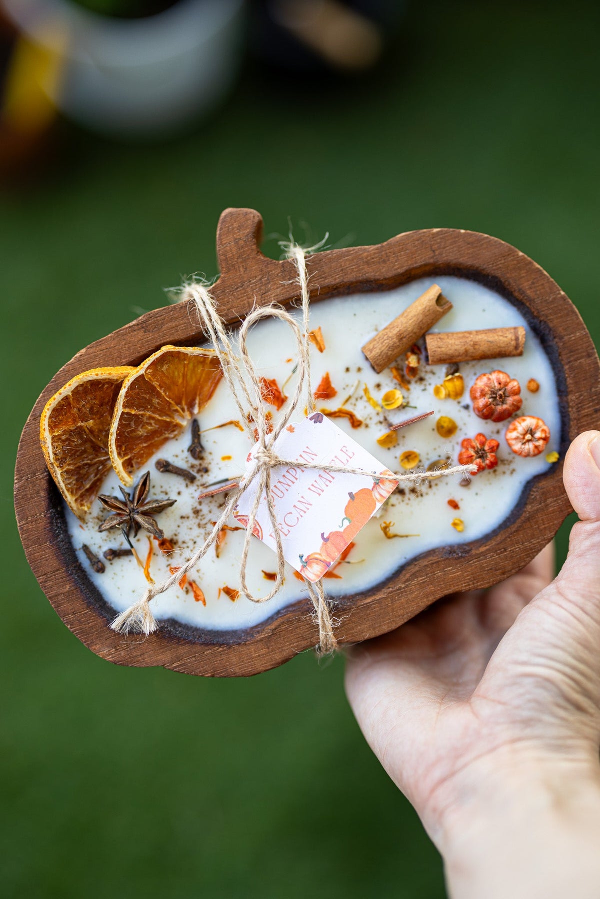 Decorative pumpkin-shaped soap with spices and oranges held by a hand against a blurred green background