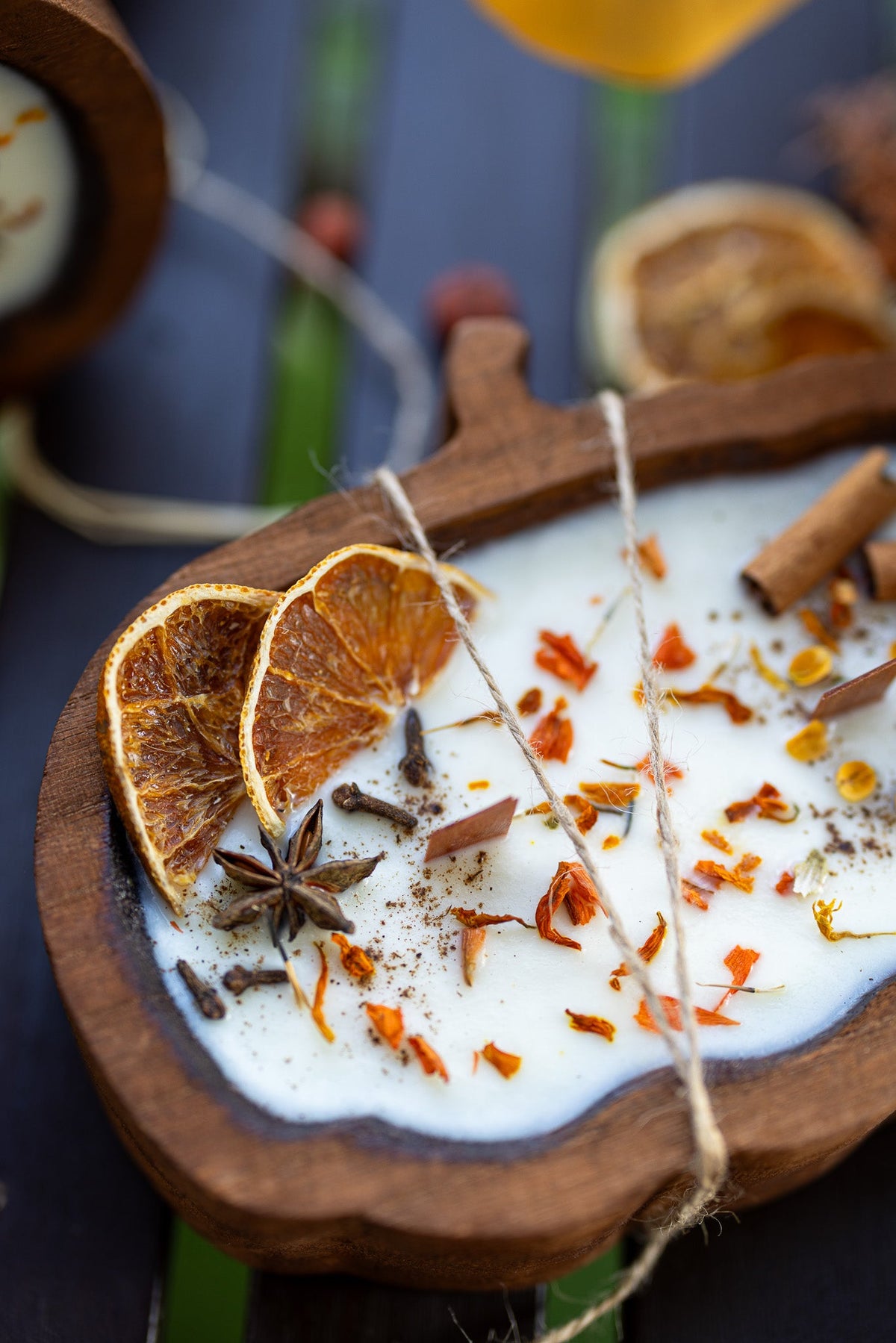A close-up of a wooden bowl candle filled with white wax and decorated with dried orange slices, star anise, cinnamon sticks, and orange flower petals on a dark rustic surface.