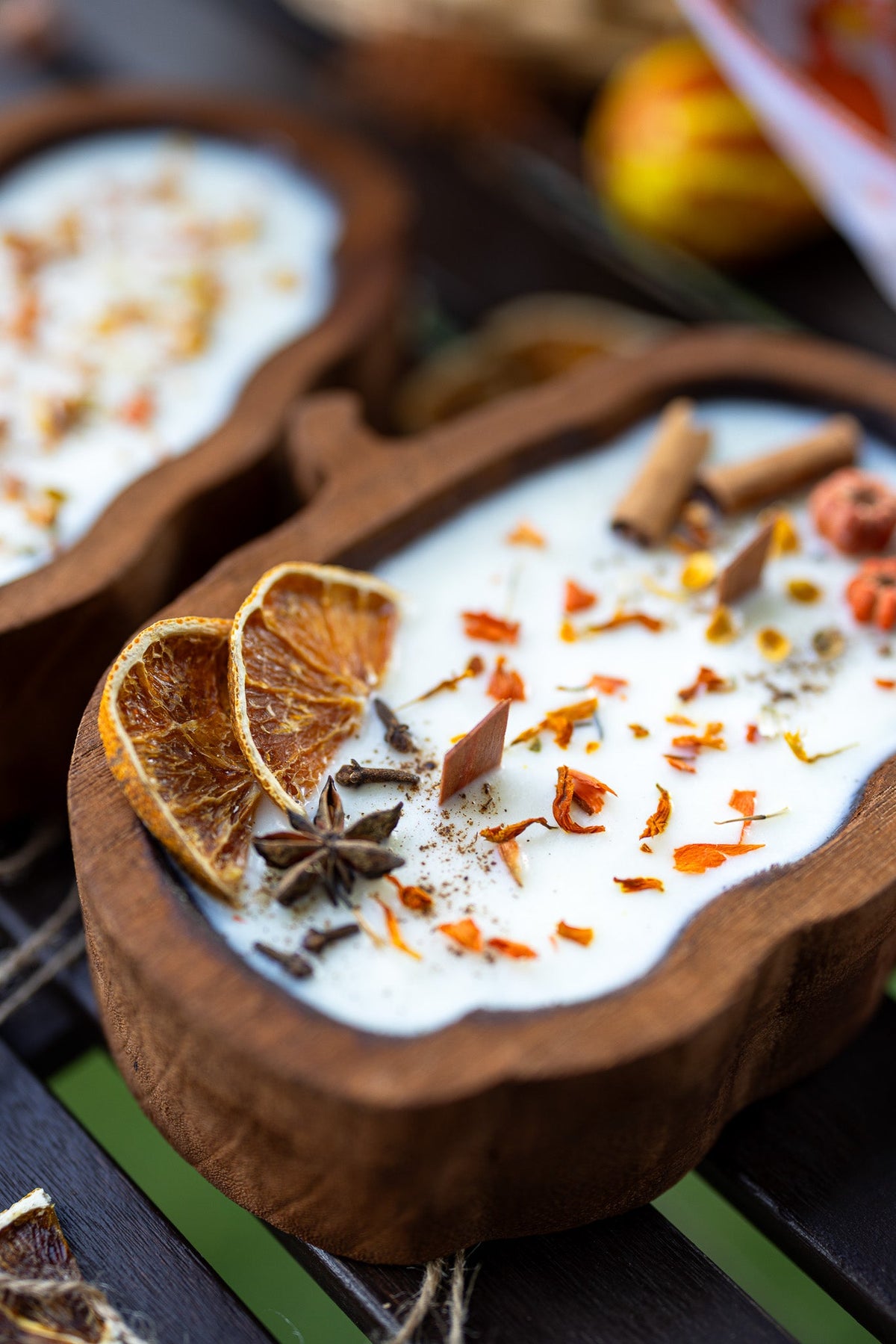 A close-up of a wooden bowl filled with a white dessert or drink, garnished with dried orange slices, cinnamon sticks, star anise, and orange flower petals, set on a dark wooden surface.