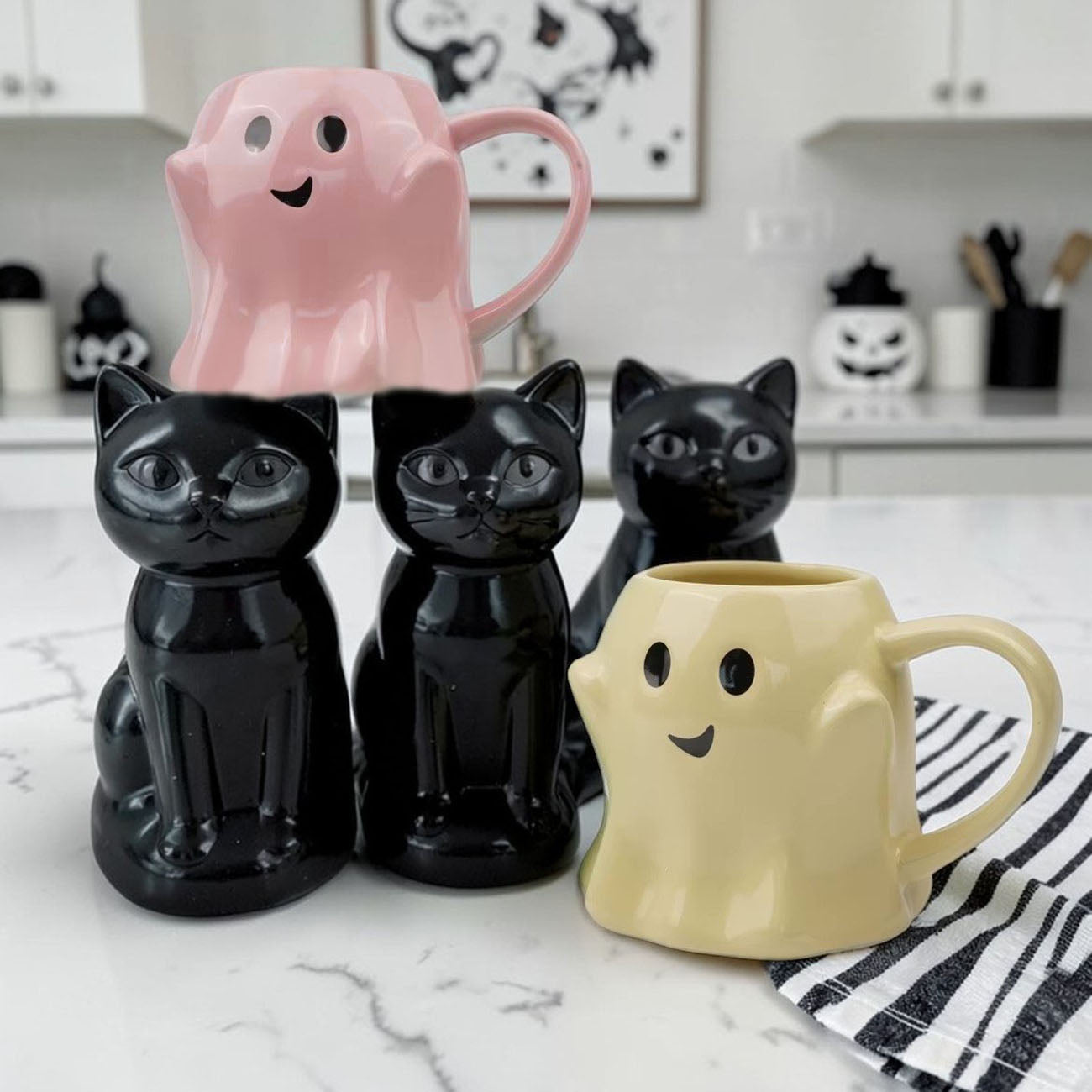 Black cat-shaped mugs and a pink ghost-shaped mug on a kitchen counter.