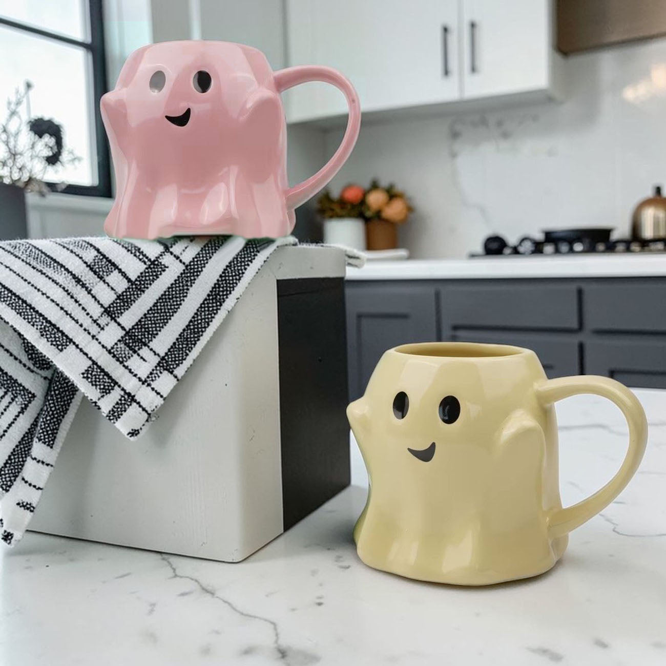 Two ghost-shaped mugs, one pink and one beige, on a kitchen counter.