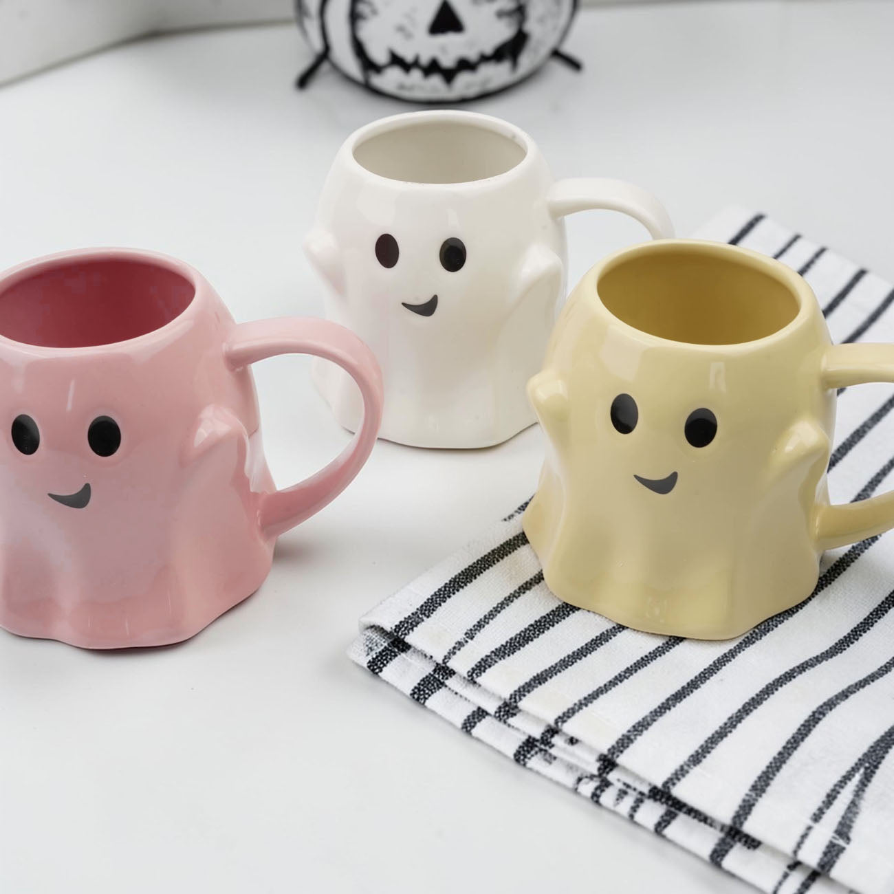 Three ghost-shaped mugs in pink, white, and yellow on a white surface with a black and white striped towel.