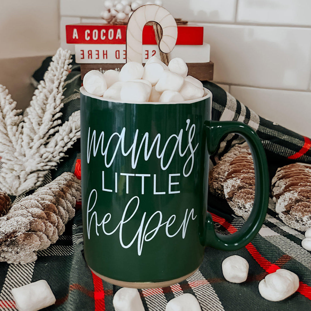 Green mug with 'mama's little helper' text, filled with marshmallows, on a cozy blanket.