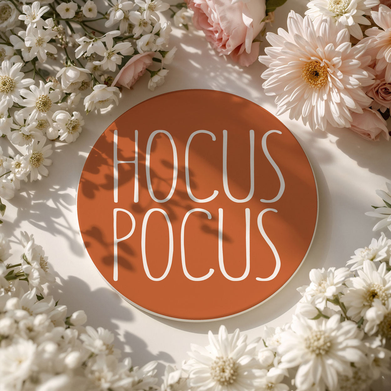 Round sign with 'HOCUS POCUS' text surrounded by flowers on a light background