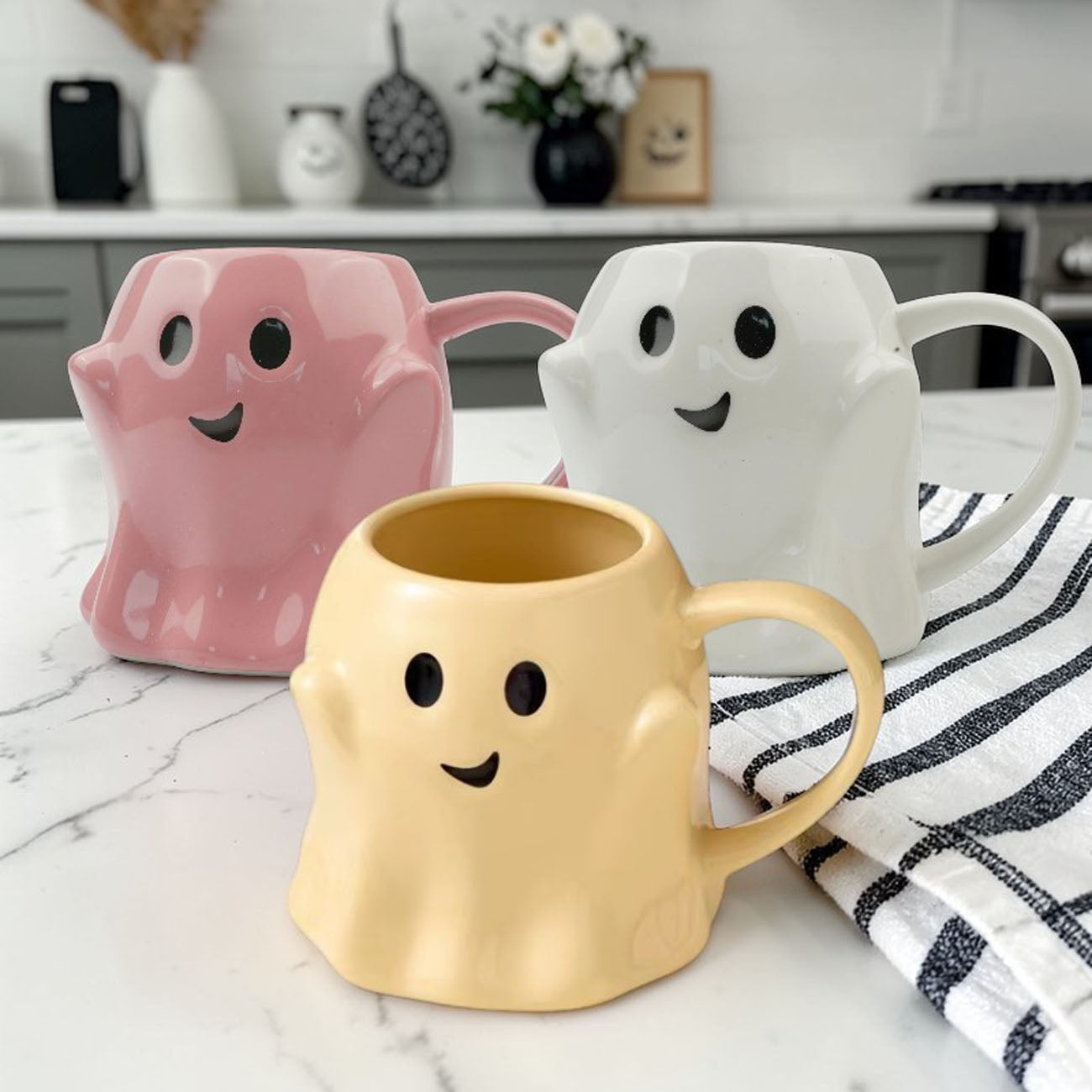 Three ghost-shaped mugs in pink, white, and yellow on a marble countertop.