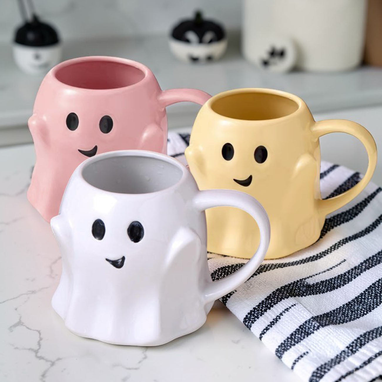Three ghost-shaped mugs in pink, white, and yellow on a marble surface.