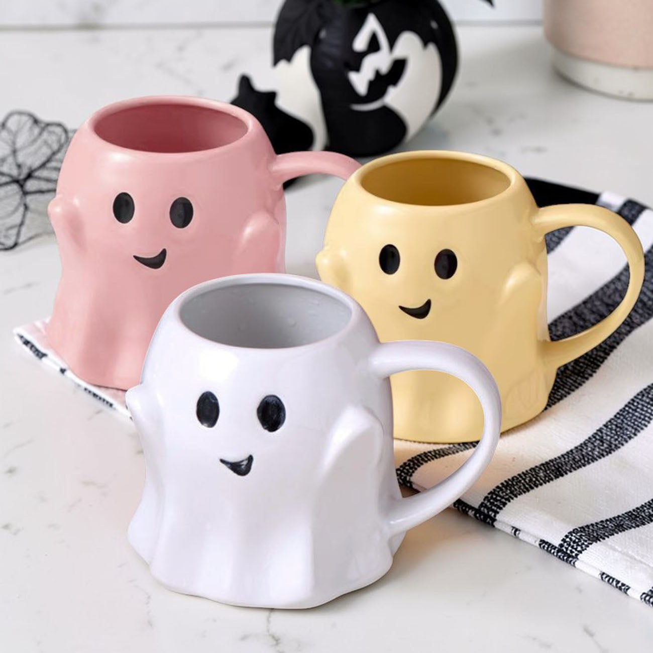 Three ghost-shaped mugs in pink, yellow, and white on a marble surface with Halloween-themed decorations.