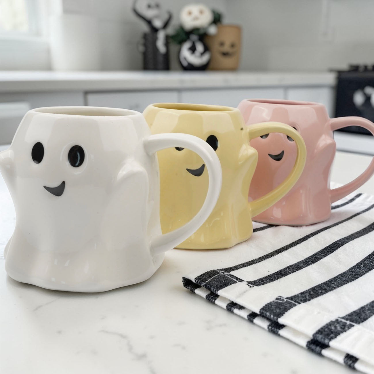 Three ghost-shaped mugs in white, yellow, and pink on a kitchen counter.