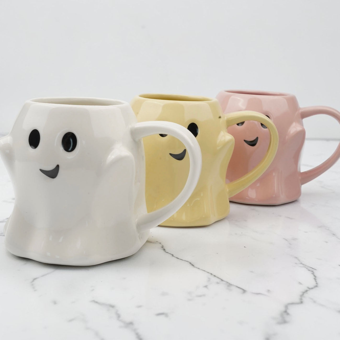 Three ceramic mugs with ghost and smiley face designs on a marble surface.