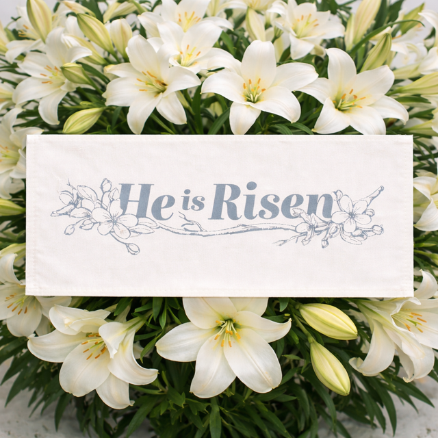 A white rectangular fabric banner or sign with the words “He is Risen” in blue script and floral line art, placed over a bouquet of white lilies.