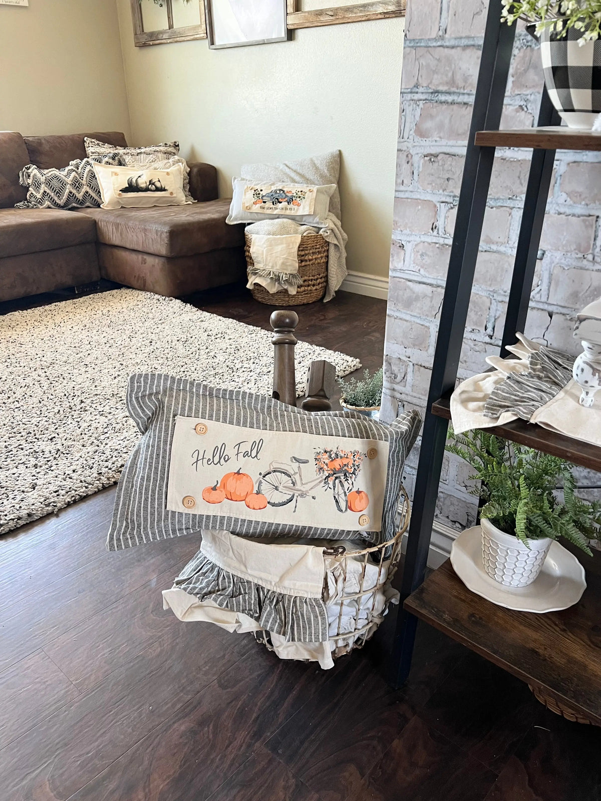 A decorative autumn-themed throw pillow with a striped fabric border and a central panel reading 'Hello Fall' with pumpkins, a bicycle, and flowers, styled in a living room next to a brick column and wooden shelf.