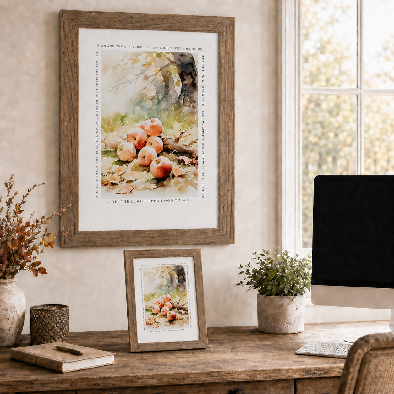 A framed art print featuring a watercolor still life of red apples on a desk in a warm, styled home office setting. A larger framed print hangs on the wall, with a smaller matching framed print resting on the wooden desk below, alongside a computer monitor, plants, and decorative items.