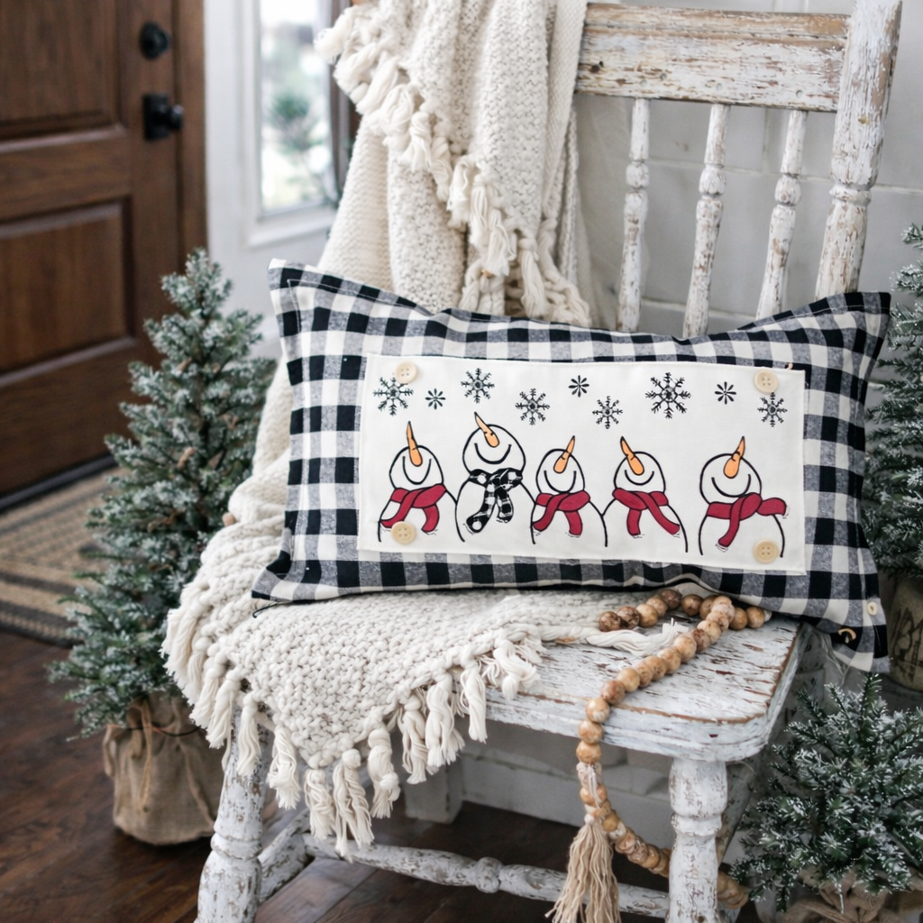 A rectangular holiday throw pillow with a black and white buffalo plaid border and a white center panel featuring five smiling snowmen, styled on a distressed white wooden chair in a cozy holiday setting.