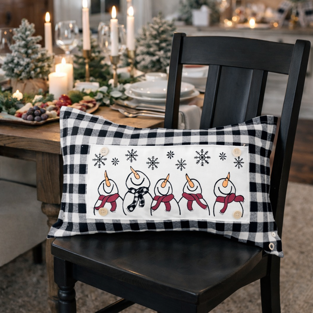 A rectangular holiday throw pillow with a black and white buffalo plaid border and a white center panel featuring five snowmen and snowflakes, displayed on a black wooden chair in a warmly lit dining room setting.