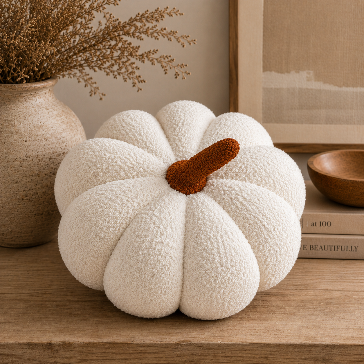 A textured white faux-fur pumpkin decoration with a brown stem, styled on a wooden shelf next to a ceramic vase with dried branches and a book.