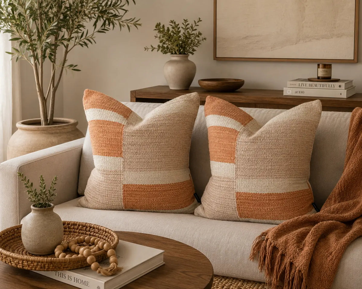 Two square woven throw pillows with horizontal stripes in beige, cream, and terracotta orange, styled on a light beige sofa in a warm, modern living room setting.