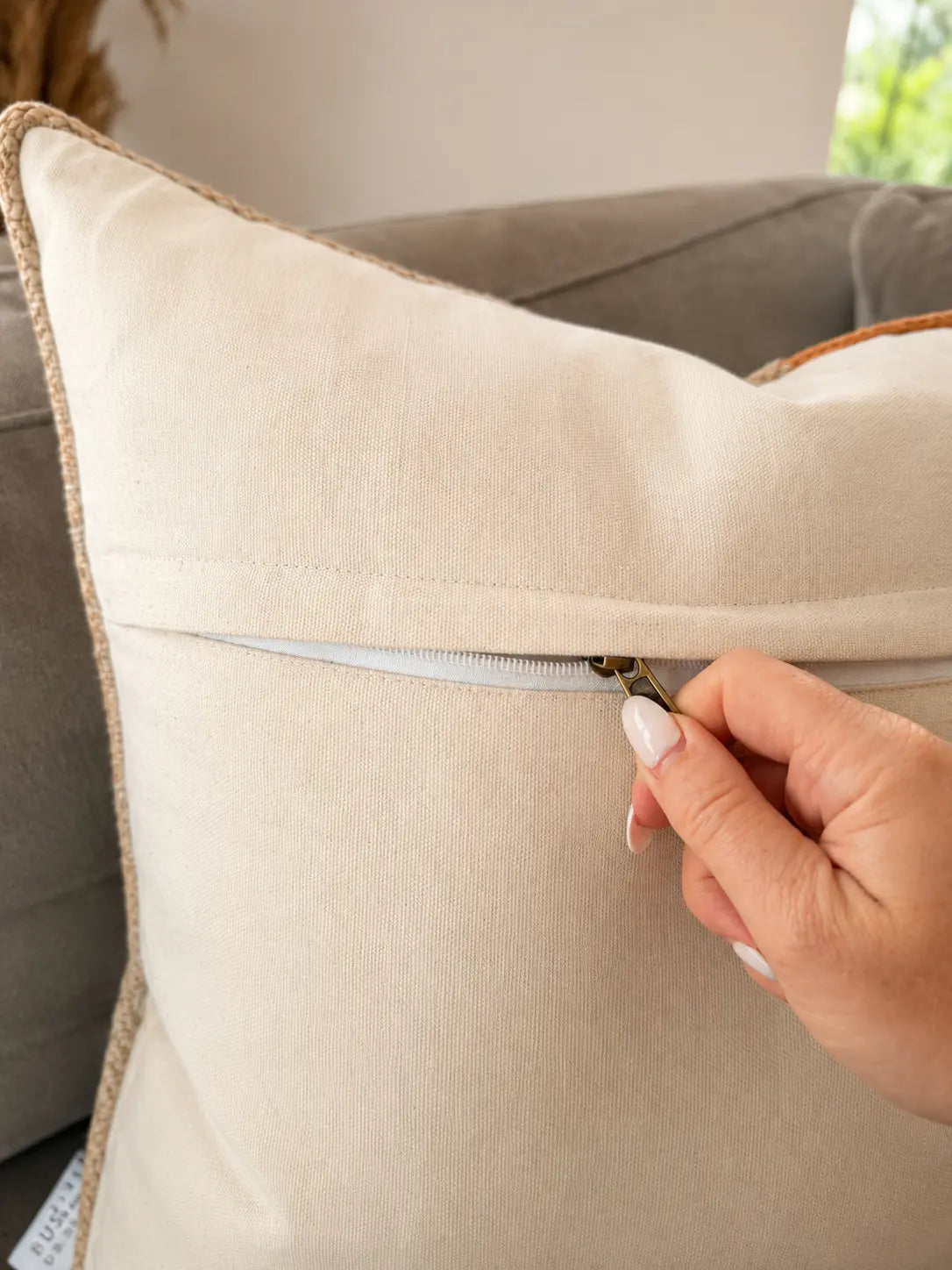 A close-up of a beige throw pillow with a zipper pocket, being opened by a hand with white manicured nails. The pillow features a natural jute trim along the edge and is set against a gray sofa background.